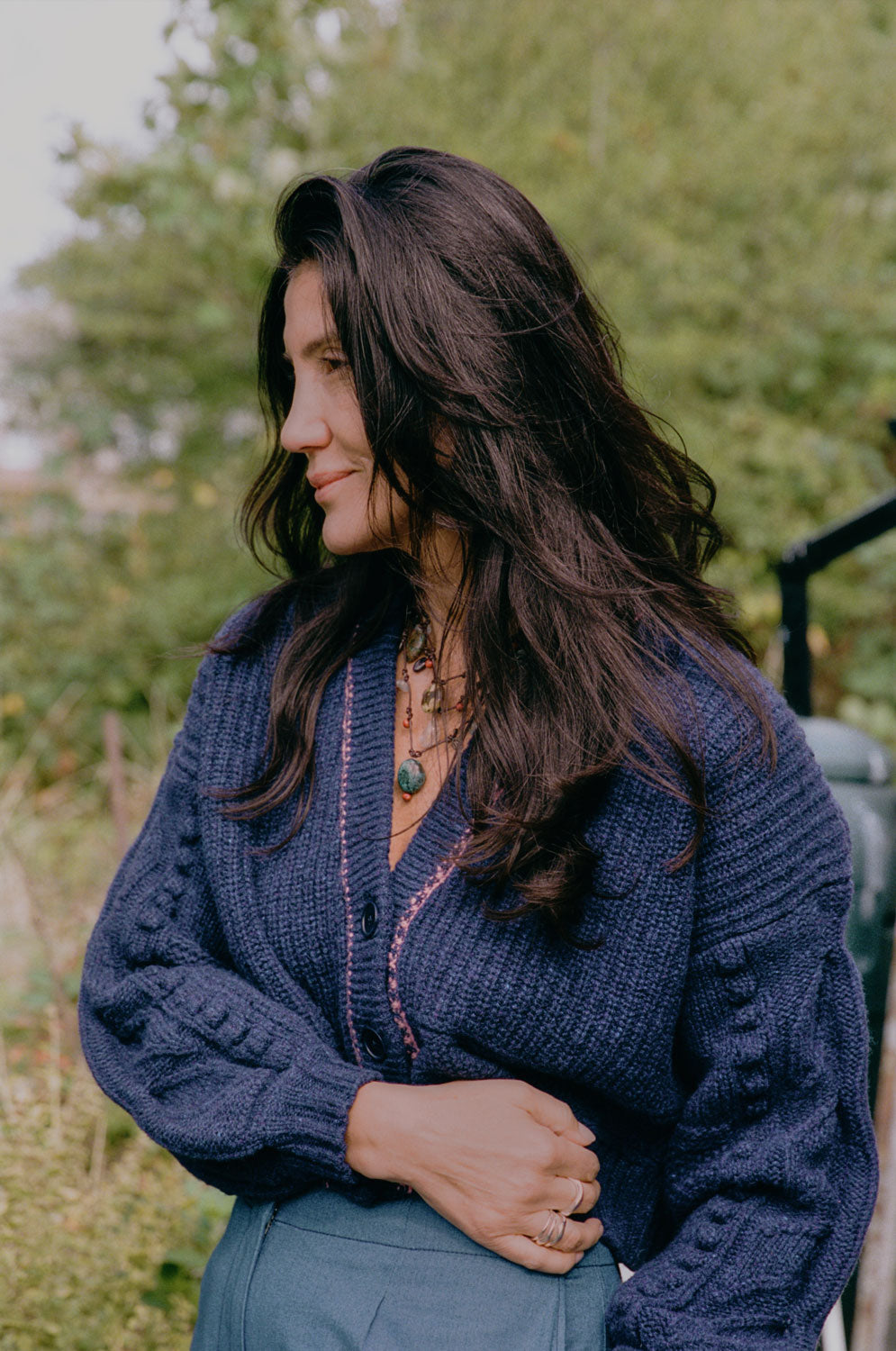 A woman stands side on in front of white brambles, her arms folded looking towards us. She wears Saywood's Amelia Wide Leg Trousers in pink corduroy with the Lela Colourblock Shirt in pink and orange. Her hair is in plaited pigtails.
