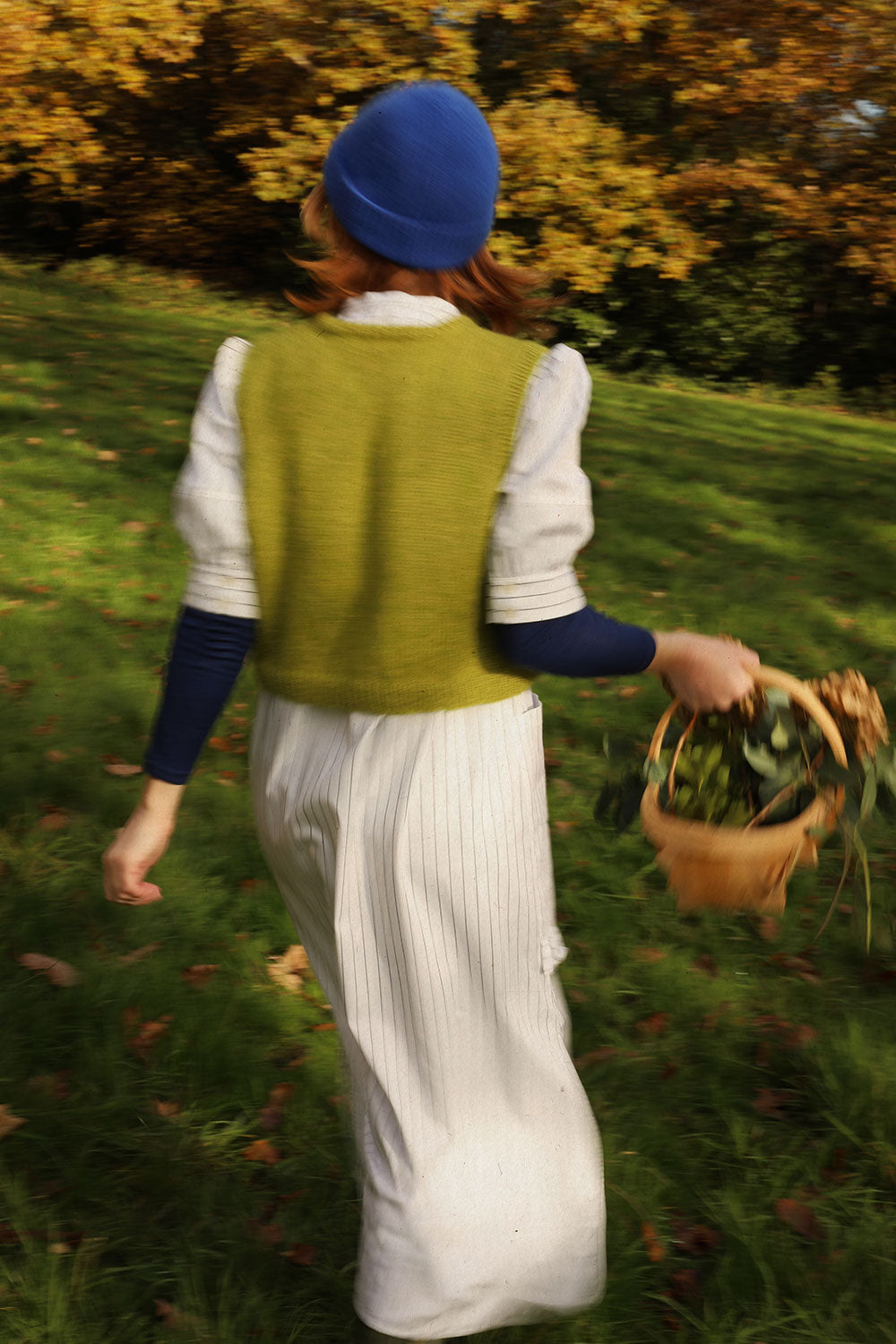A woman is walking in a field with her back to the camera. She holds a trugg of flowes, and is wearing Saywood's white pinstripe Rosa Puff Sleeve Dress with a green knit sweater vest, navy long sleeve underlayer and cobalt blue beanie hat.