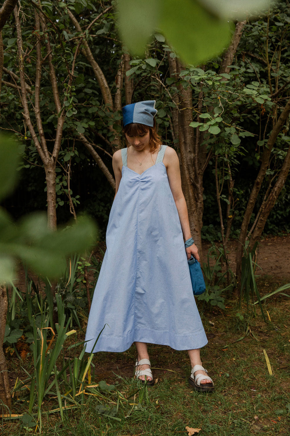 Maya Cami Dress in pale blue recycled cotton, worn with headscarf, full length model in a woodland setting. By Saywood. Made in London.