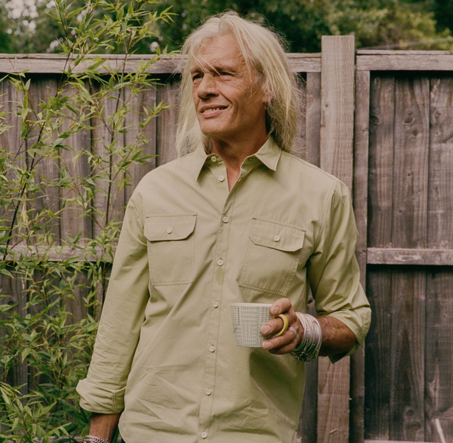 A man with long blonde grey hair stands in the garden in front of a plant bed and a wooden fence. He is holding a cup of tea. He wears Saywood's Men's Eddy Utility Shirt with utility style pockets in olive, worn with silver metal bangles.