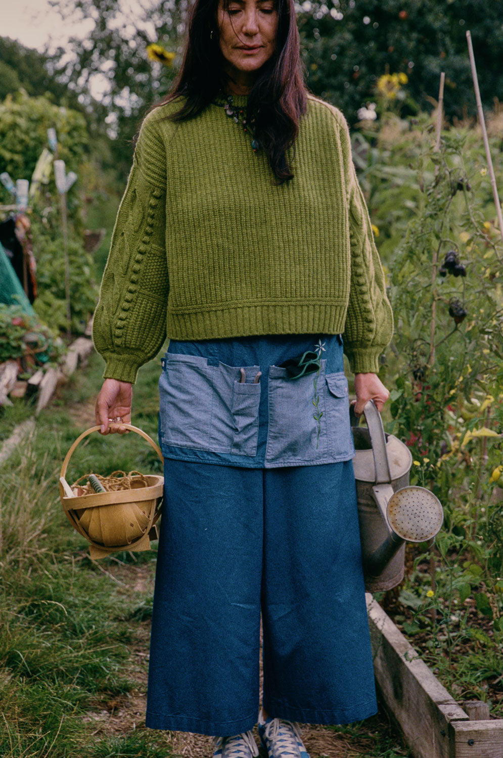 Adams boxy sweater in moss green lambswool with leaf sleeve detail, styled with wide leg denim trousers and garden apron, outdoor lifestyle shot