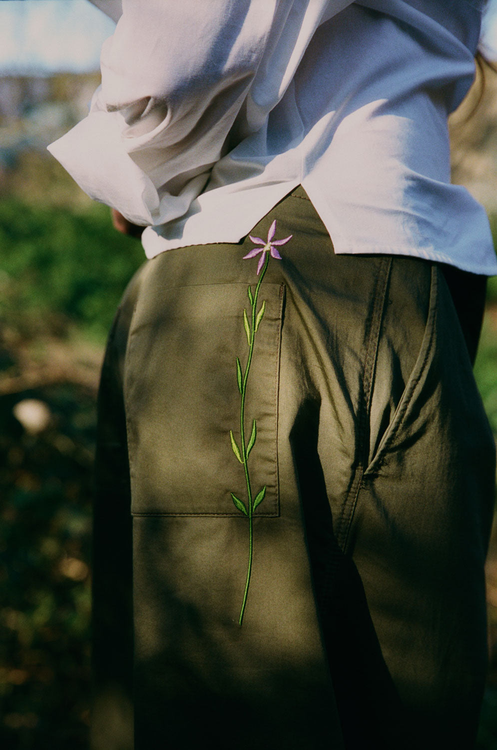 Detail view of Lela white recycled cotton shirt showing side split hem, paired with khaki Amelia trousers featuring signature embroidered flower detail by Saywood